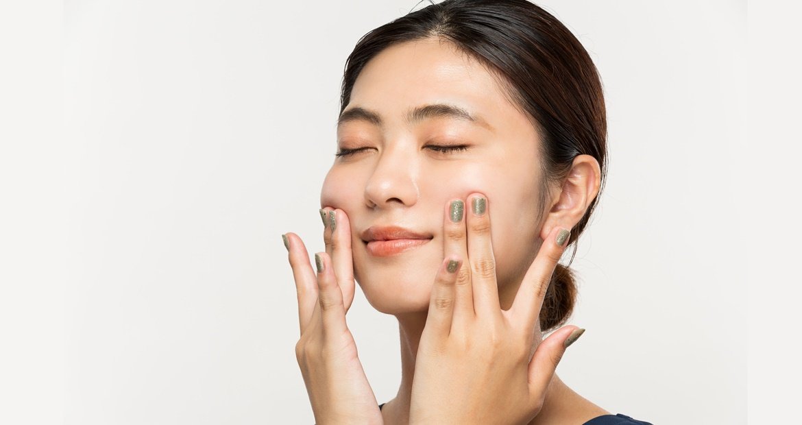 Jovem mulher com traços asiáticos toca ambos os lados de seu rosto que possui uma pele lisa, uniforme e saudável